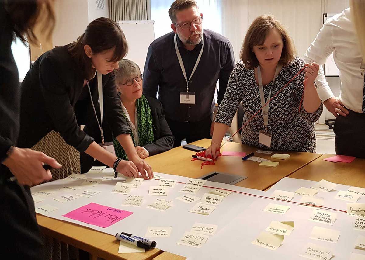 Eine Gruppe steht versammelt um einen Tisch und verbindet verschiedene Post-Its mit rotem Faden (Team & Leadership Coaching)